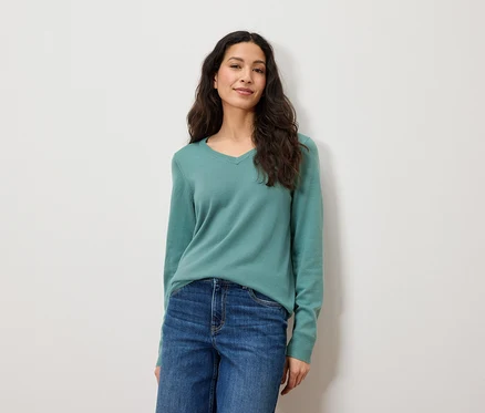 Eine Frau mit langen braunen Haaren posiert vor einem weißen Hintergrund. Sie trägt blaue Jeans und einen grünen Pullover.