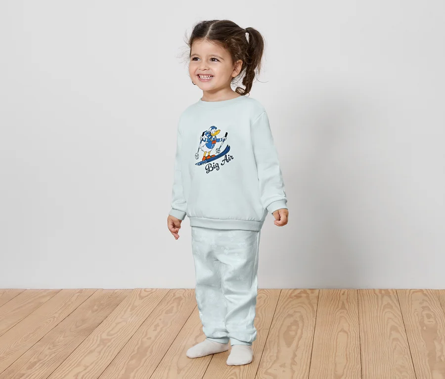 Lächelndes kleines Mädchen in hellblauem Set mit einem Cartoon-Charakter-Print auf einem Snowboard.