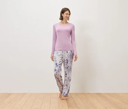 Frau steht barfuß auf einem Holzboden, trägt einen lila Pullover und eine bedruckte Hose.