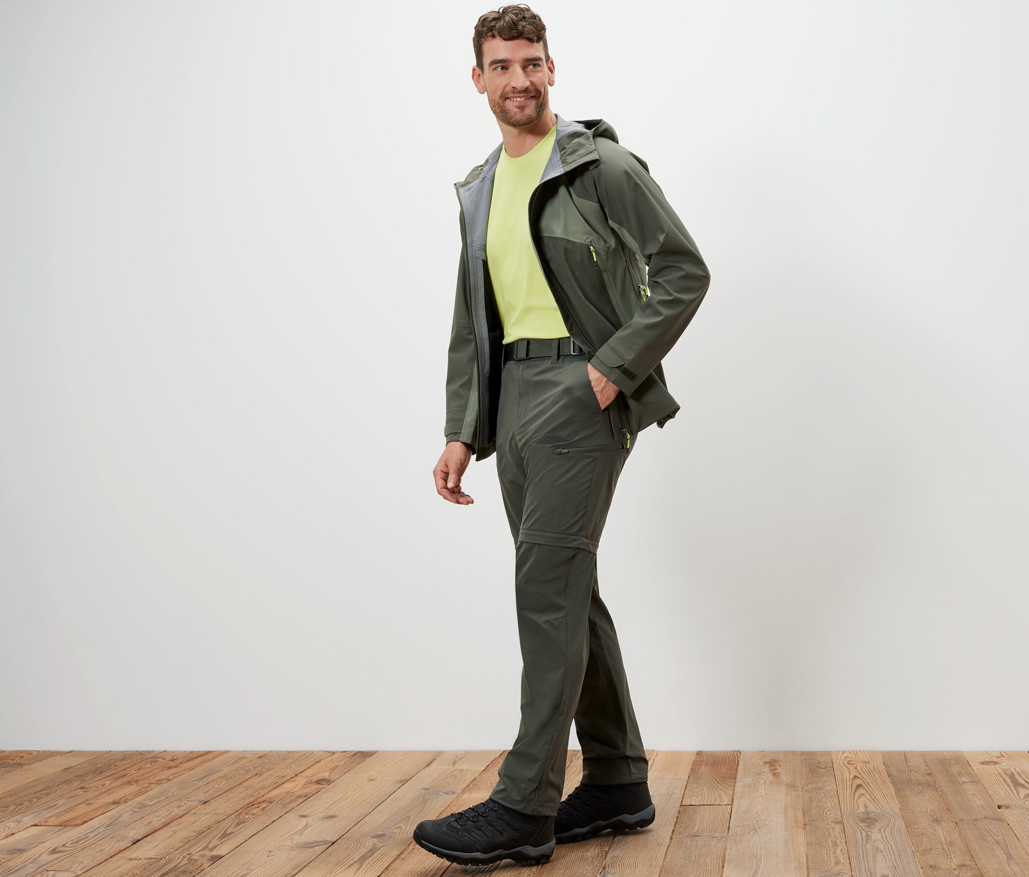 Mann posiert in Khaki 2-in-1-Funktionshose und Jacke auf Holzboden.