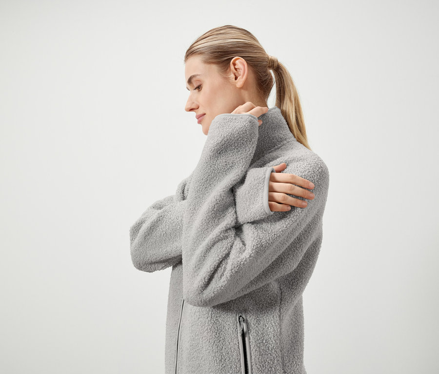 Profil einer Frau mit blonden, zu einem Pferdeschwanz zusammengebundenen Haaren, die eine graue, flauschige Jacke mit Reißverschluss trägt und vor einem weißen Hintergrund steht.