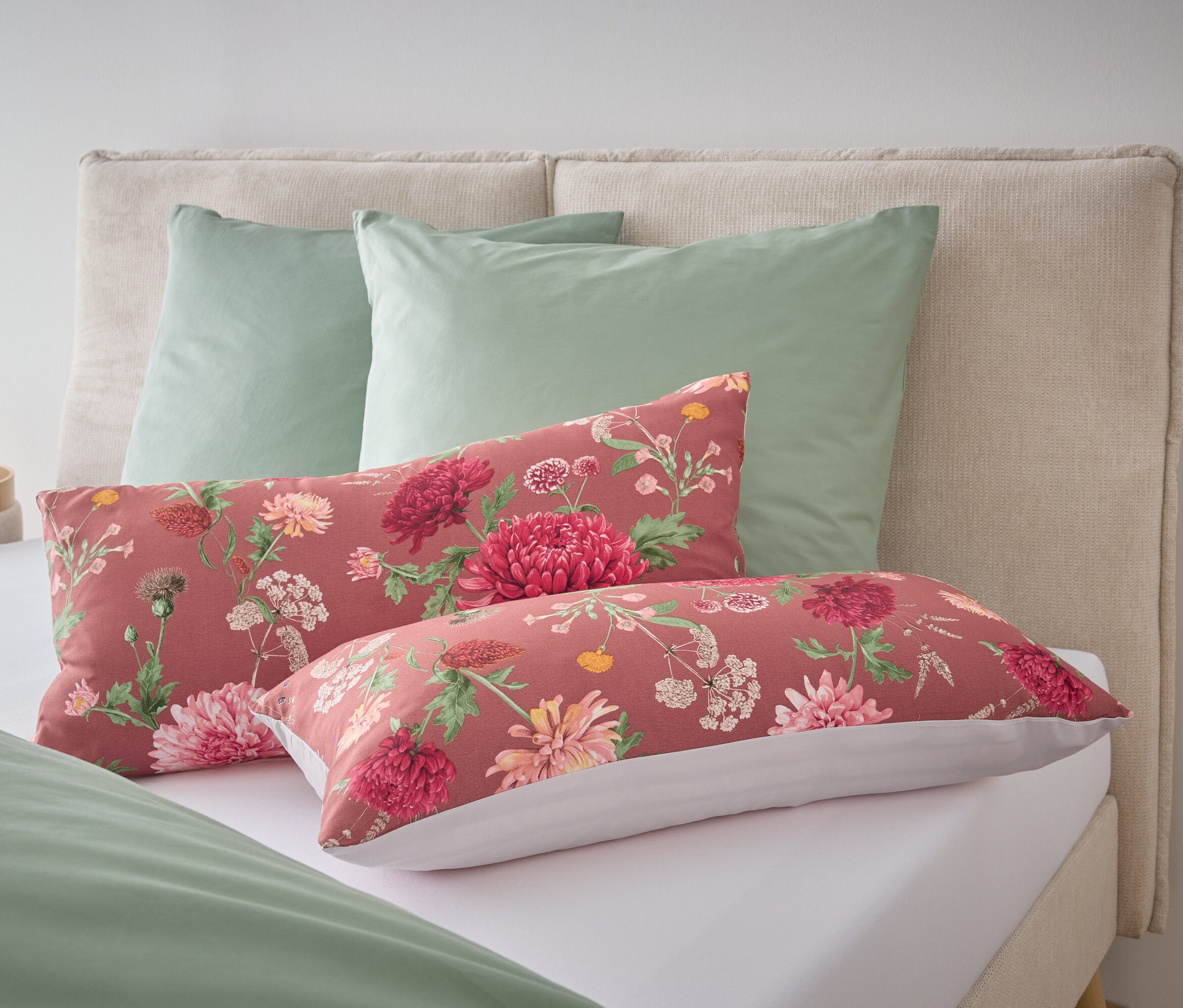 Auf einem Bett liegen zwei grüne Kissen und zwei längliche Kissen mit Blumenmuster.