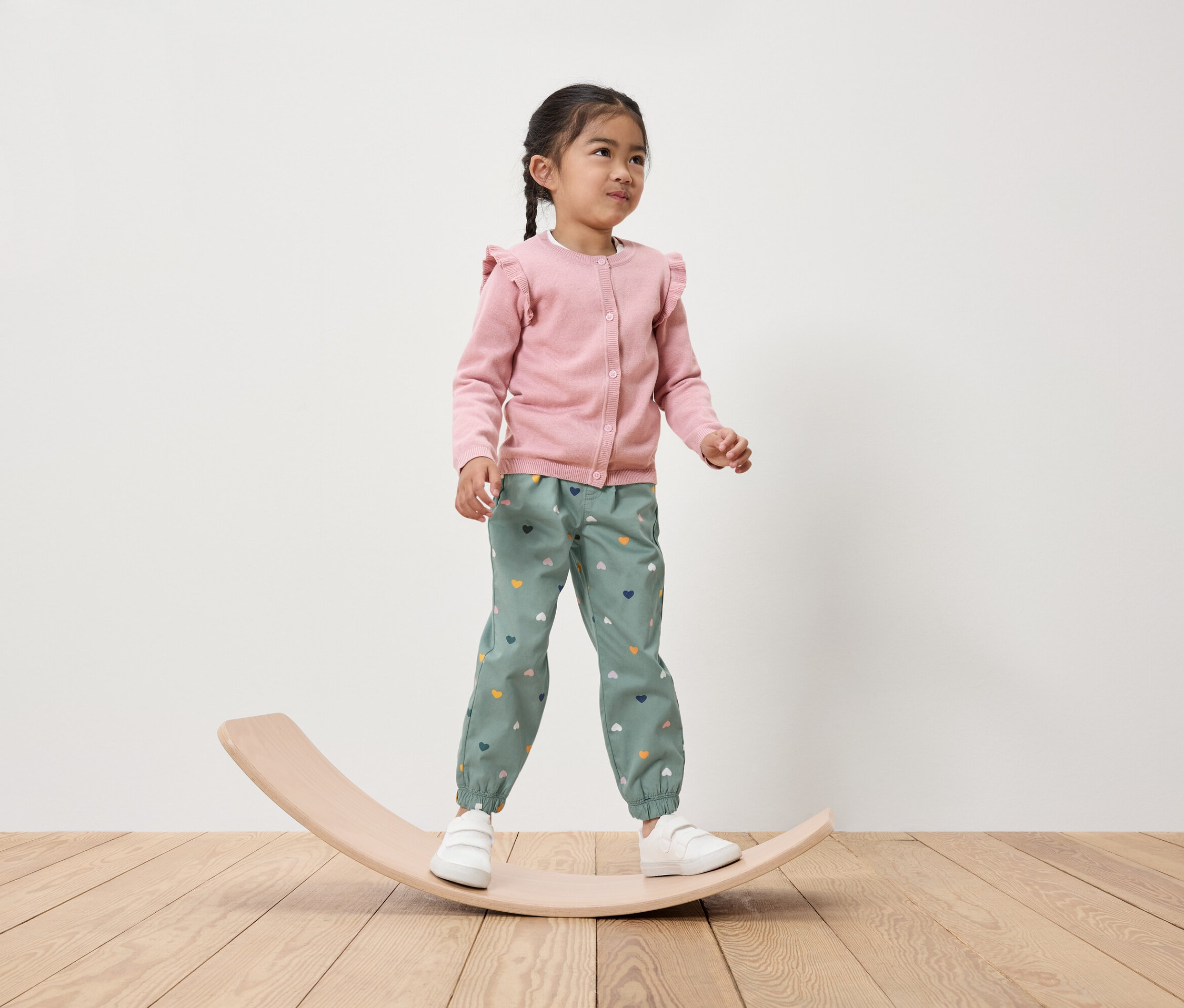 Mädchen steht auf einem hölzernen Balancebrett, trägt einen rosa Cardigan und grüne Hosen mit Herzen.