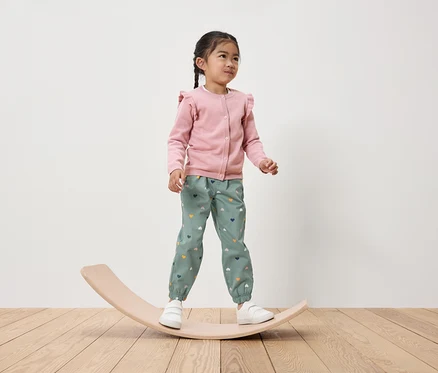 Mädchen steht auf einem hölzernen Balancebrett, trägt einen rosa Cardigan und grüne Hosen mit Herzen.