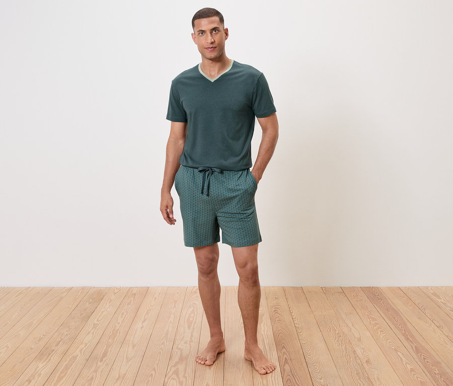Mann steht barfuß auf Holzboden und trägt einen grünen Schlafanzug mit kurzen Ärmeln und Shorts.