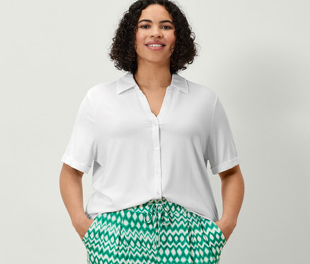 Eine Frau steht mit den Händen in den Taschen einer bedruckten 3/4-Jerseyhose und einem weißen Blusenshirt mit Knopfleiste.