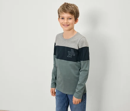 Ein Junge in einem langärmligen Shirt mit grauem Oberteil, dunkelblauem Streifen mit LA-Logo und grünem Unterteil steht vor einer weißen Wand.