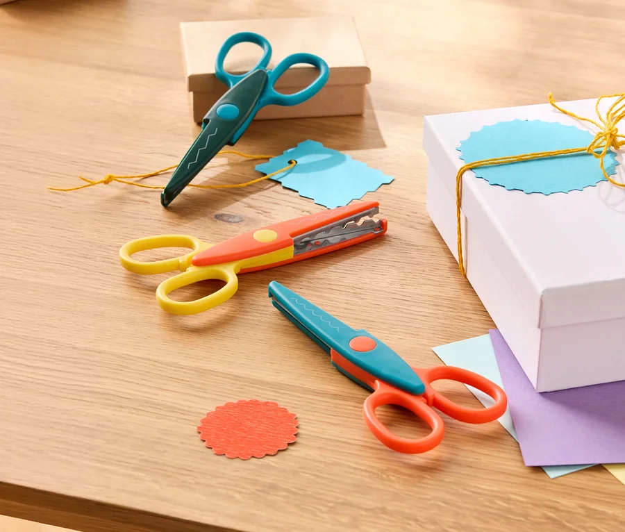 Drei Zierscheren in blau-grün, gelb-orange und blau-orange liegen auf einem Holztisch neben Geschenkboxen und farbigem Papier.