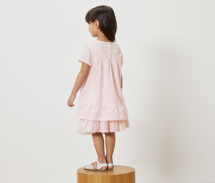Mädchen steht auf einem Hocker und trägt ein rosa-weiß gestreiftes Kleid und weiße Sandalen.