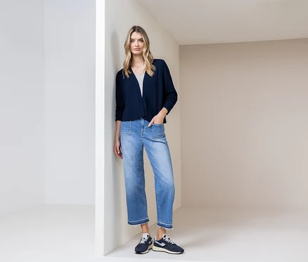 Frau lehnt an einer Wand, trägt eine marineblaue Strickjacke, blaue Jeans und marineblaue Schuhe.