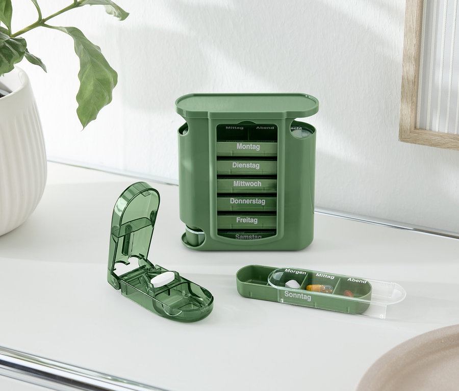 Auf einem Tisch stehen ein grüner Pillensortierer, ein Tablettenteiler und eine Pillendose neben einer Topfpflanze.