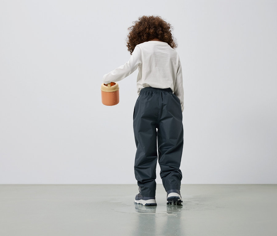Kind von hinten gesehen, hält einen orangefarbenen Eimer in der Hand, trägt eine Hose in Anthrazit und steht in einer Wasserpfütze.