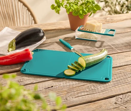 Auf einem Holztisch liegen ein roter Pfeffer, eine Aubergine, ein grünes Schneidebrett mit einem Messer und einer geschnittenen Zucchini, eine Käsereibe mit Käse und ein Topf mit einer Pflanze.