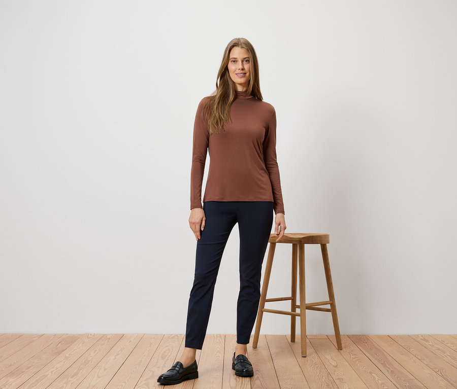 Frau in braunem Rollkragenshirt und blauen Hosen posiert neben einem Hocker auf einem Holzboden.