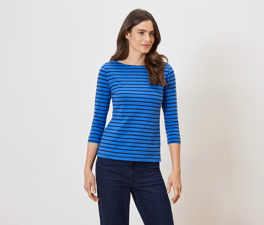 Eine Frau mit langen, gewellten, braunen Haaren trägt ein blaues, horizontal gestreiftes Oberteil und eine dunkle Jeans.