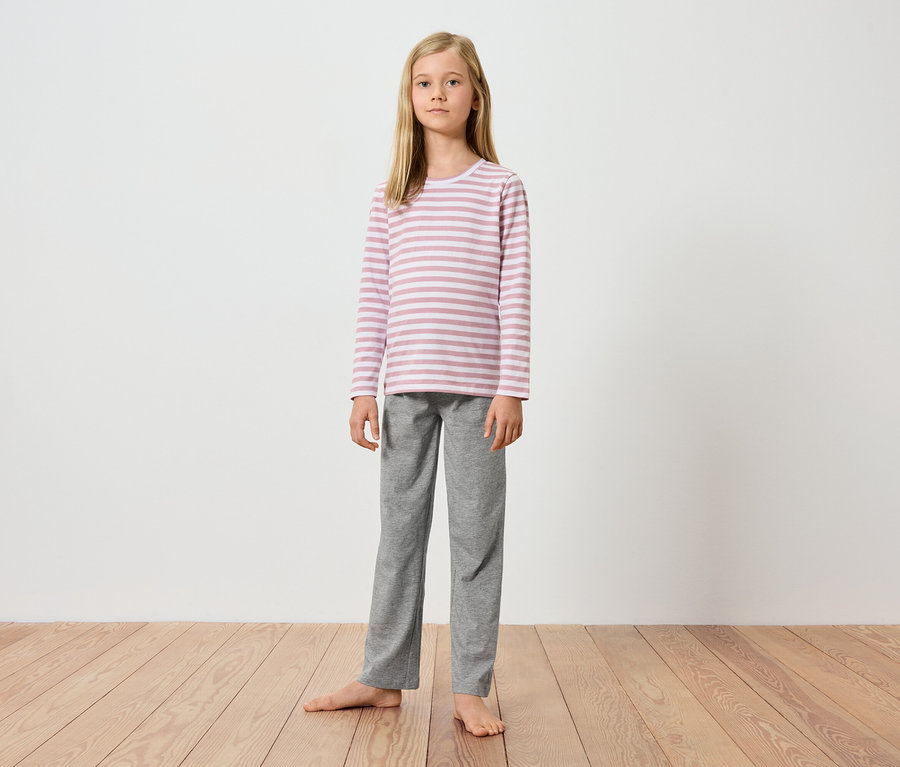 Ein junges Mädchen steht barfuß auf einem Holzboden vor einer weißen Wand. Sie trägt ein langärmeliges, rosa-weiß gestreiftes Oberteil und eine graue Hose.
