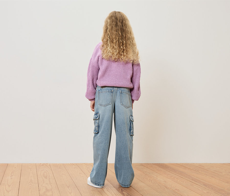 Rückansicht eines Mädchens mit blonden Locken, das einen lila Pullover und hellblaue Jeans mit Seitentaschen trägt.