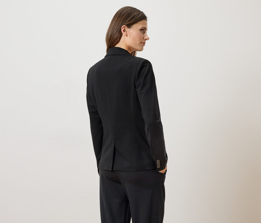 Frau von hinten in schwarzem Blazer und Hose, eine Hand in der Hosentasche, Blick über die Schulter.