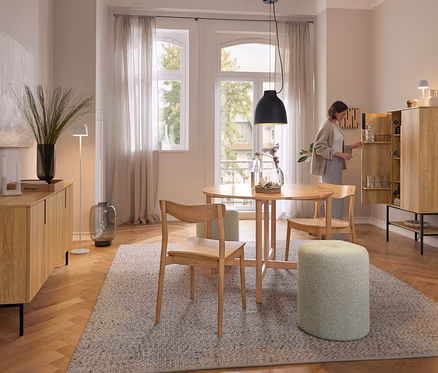 Eine Frau öffnet einen hellen Holzschrank in einem Esszimmer. Über einem runden Holztisch hängt die schwarze Eglo Pendelleuchte »Camasca«. Auf dem Schrank steht die weiße Eglo LED-Akku-Tischleuchte »Mannera«.