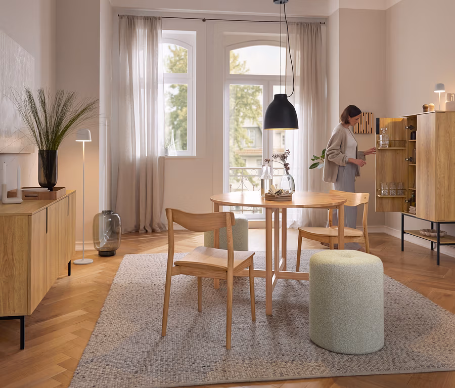 Eine Frau öffnet einen hellen Holzschrank in einem Esszimmer. Über einem runden Holztisch hängt die schwarze Eglo Pendelleuchte »Camasca«. Auf dem Schrank steht die weiße Eglo LED-Akku-Tischleuchte »Mannera«.
