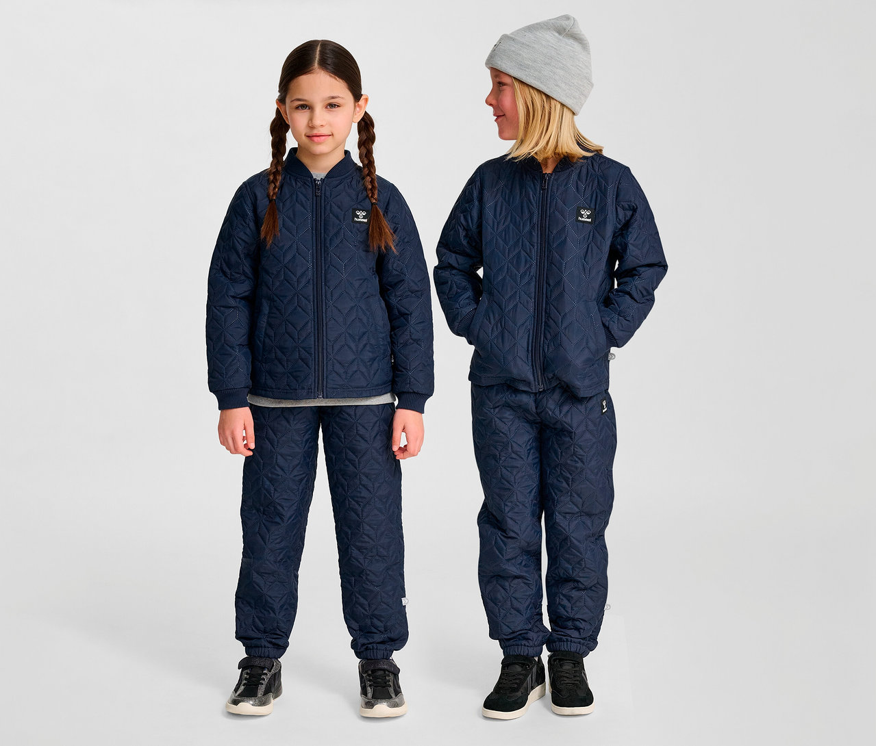 Zwei Kinder stehen in blauen, gesteppten hummel® »HMLSOBI Thermo-Sets« mit Jacke und Hose. Ein Kind trägt eine graue Mütze.