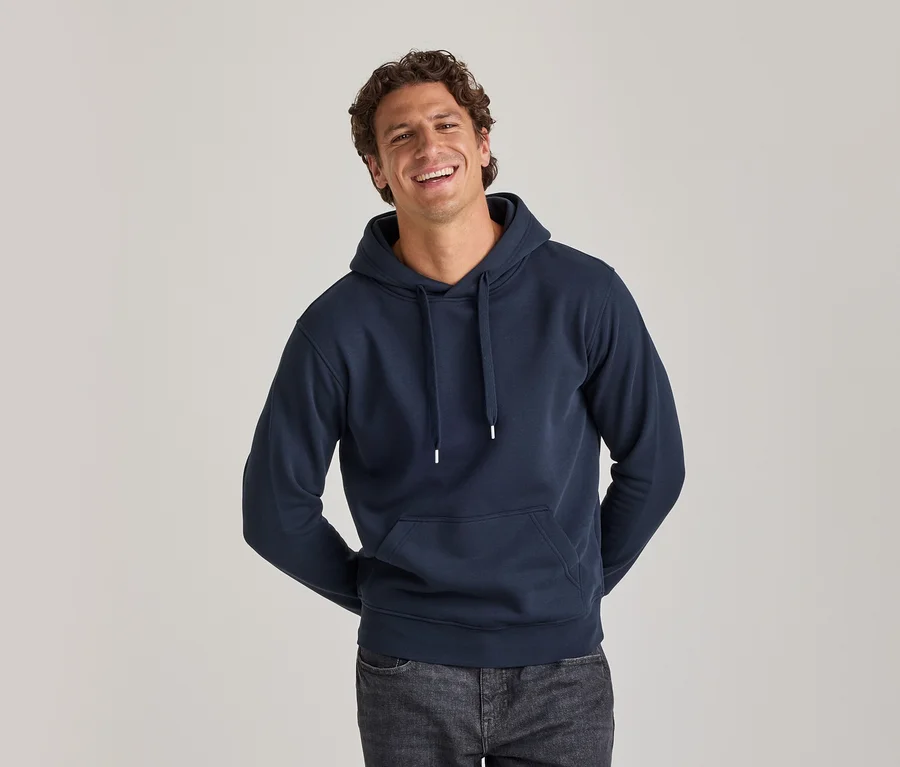Lächelnder Mann in einem marineblauen Hoodie mit weißen Kordeln und dunklen Jeans, die Hände hinter dem Rücken.