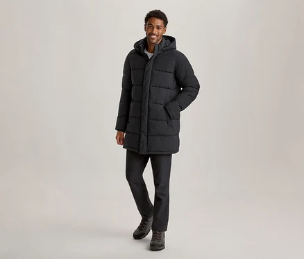 Lächelnder Mann in einer schwarzen, kapuzenbesetzten Daunenjacke, dunklen Hosen und Wanderschuhen, der in die Kamera blickt.