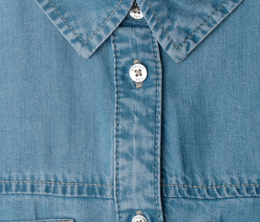 Nahaufnahme eines hellblauen Street One Jeanskleides mit Kragen und Knopfleiste. Die weißen Knöpfe tragen die Aufschrift 'Street One'.