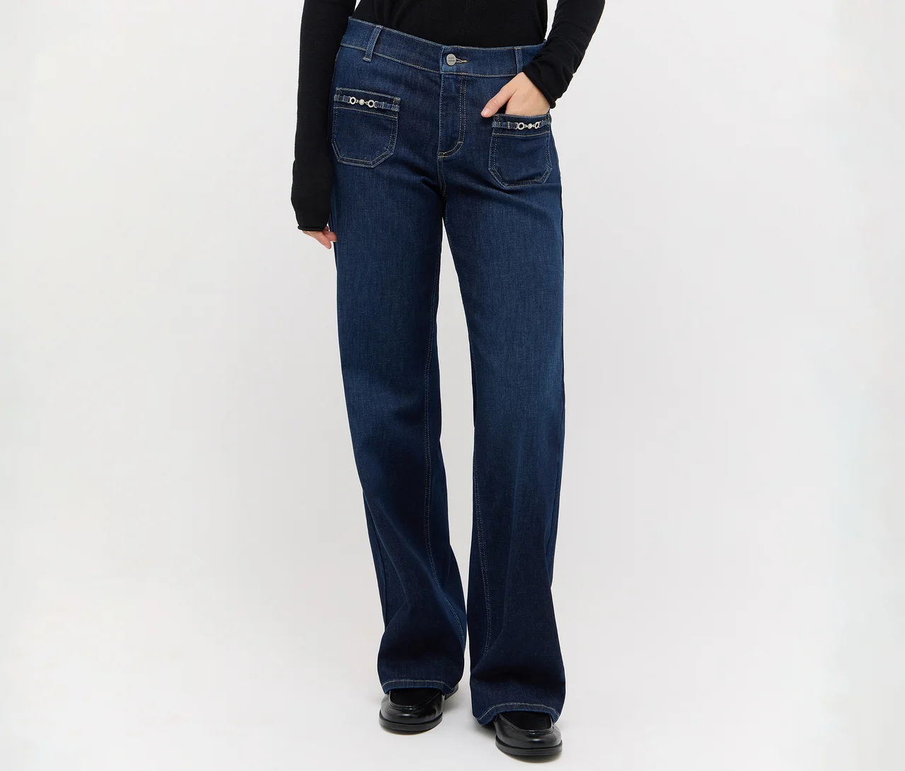 Person trägt dunkelblaue Jeans mit weitem Bein, dekorativen Vordertaschen, ein schwarzes Langarmshirt und schwarze Schuhe, eine Hand in der Tasche.