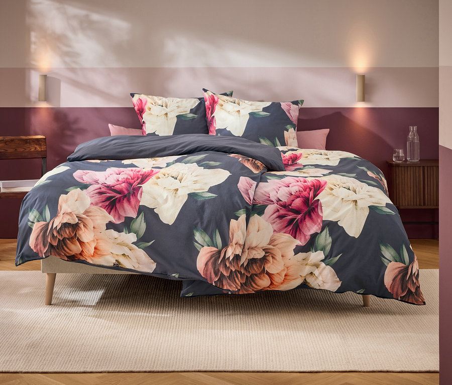 Ein Bett mit dunkelgrauer Bettwäsche und großen rosa, weißen und orangefarbenen Blumenmustern steht in einem Schlafzimmer mit zweifarbiger Wand und Holzmöbeln.