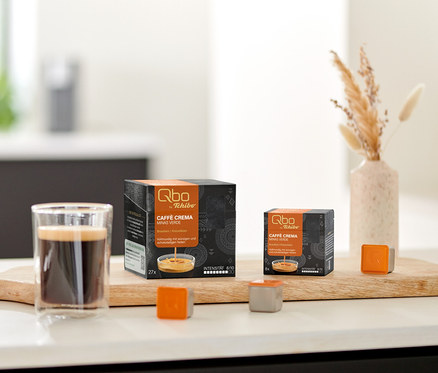 Zwei Qbo Caffè MINAS VERDE Kapselpackungen, ein Glas Kaffee und lose Kapseln auf einem Holzbrett auf der Küchenzeile, daneben Kekse und Trockenblumen.