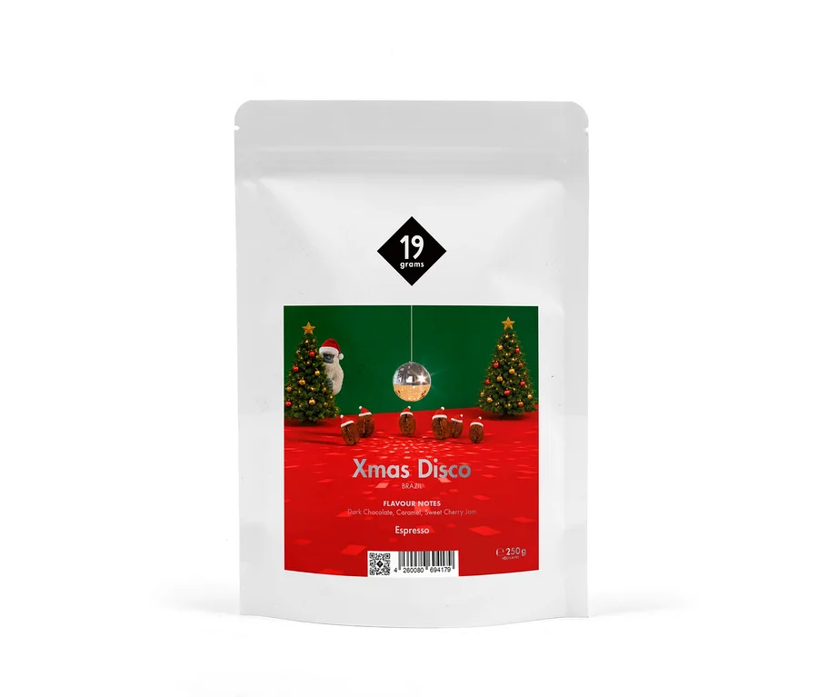 Weißer Kaffeebeutel mit rotem Etikett, das eine Weihnachts-Disco-Szene zeigt: zwei geschmückte Christbäume, ein Faultier mit Weihnachtsmütze, eine Discokugel und kleine braune Kuchen auf rotem Boden vor einer grünen Wand. Das Etikett trägt das „19 grams“-Logo, „Xmas Disco Brazil“, „Flavour Notes: Dark Chocolate, Caramel, Sweet Cherry Jam“, „Espresso“ und „250g“.