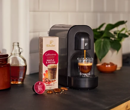 Schwarze Cafissimo now Kaffeemaschine brüht Cafissimo Flavoured Espresso Maple & Pecan in ein Glas. Daneben Kapselpackung, lose Kapsel, Nüsse, Flasche und Pflanze.