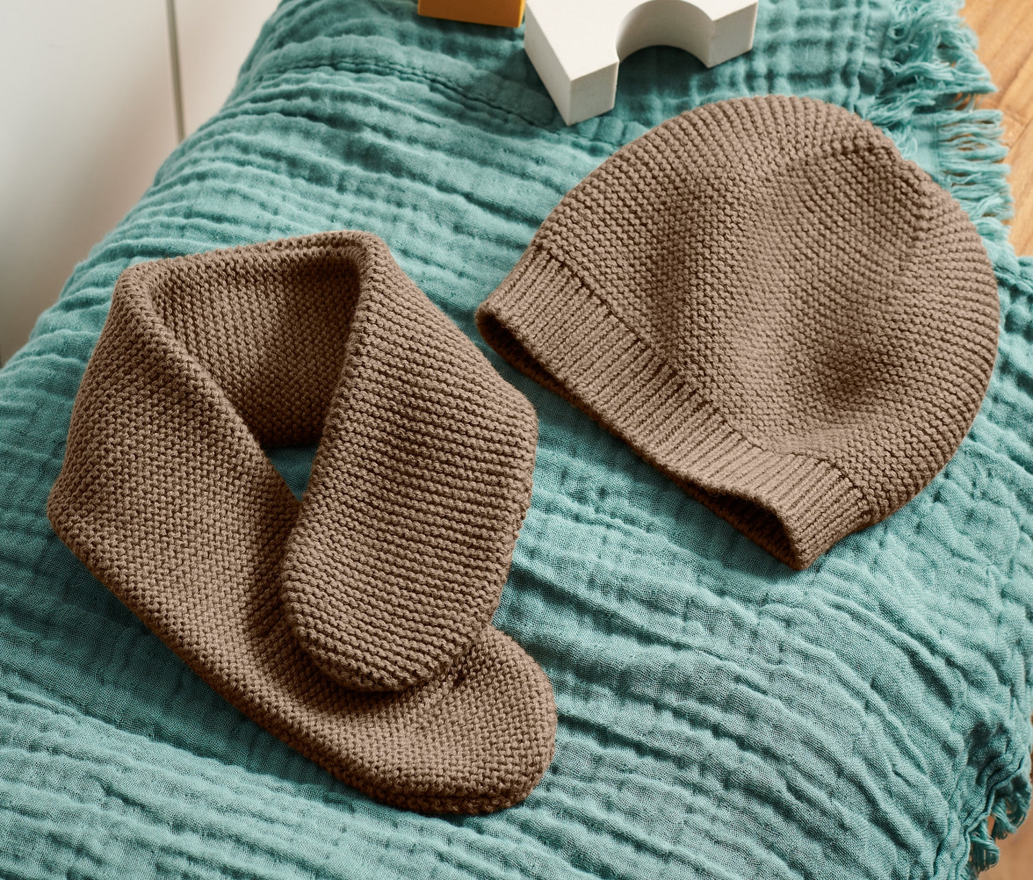 Eine braune Strickmütze und ein Strickschal liegen auf einem blaugrünen Stoff auf einem Holzhocker, neben weißen und gelben Würfeln.