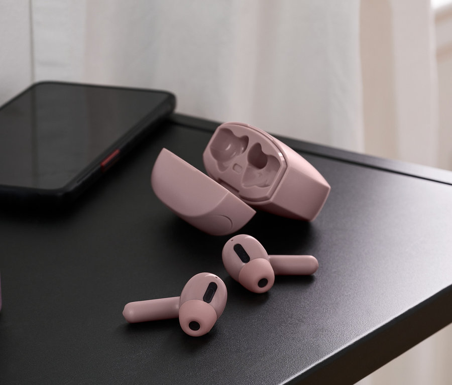 Auf einem schwarzen Tisch liegen roséfarbene TWS-In-Ear-Kopfhörer, daneben ein geöffnetes Ladecase und ein Handy. Im Hintergrund eine Vase.