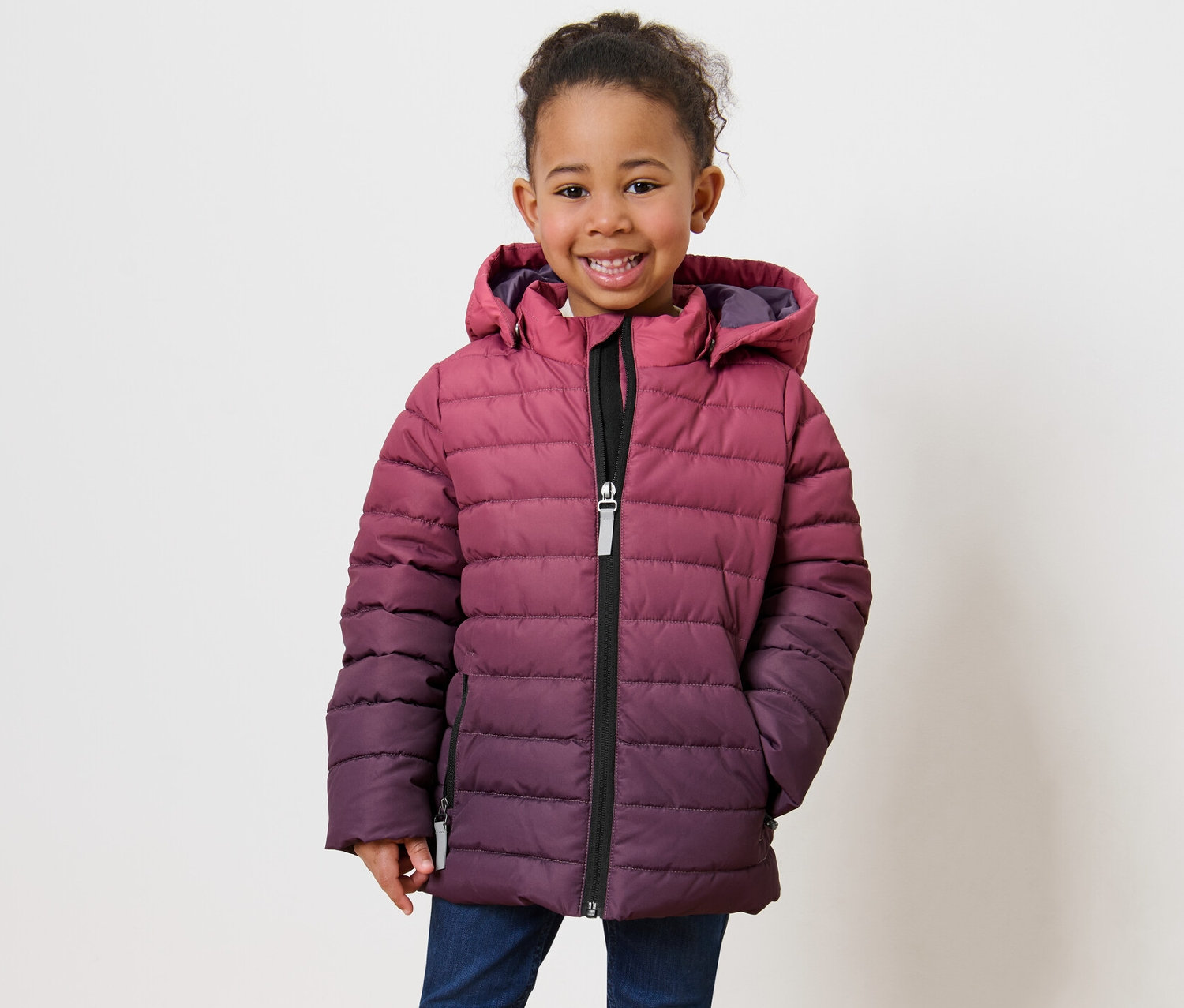 Ein lächelndes kleines Mädchen posiert in einer Kinder-Steppjacke mit Farbverlauf und Kapuze.