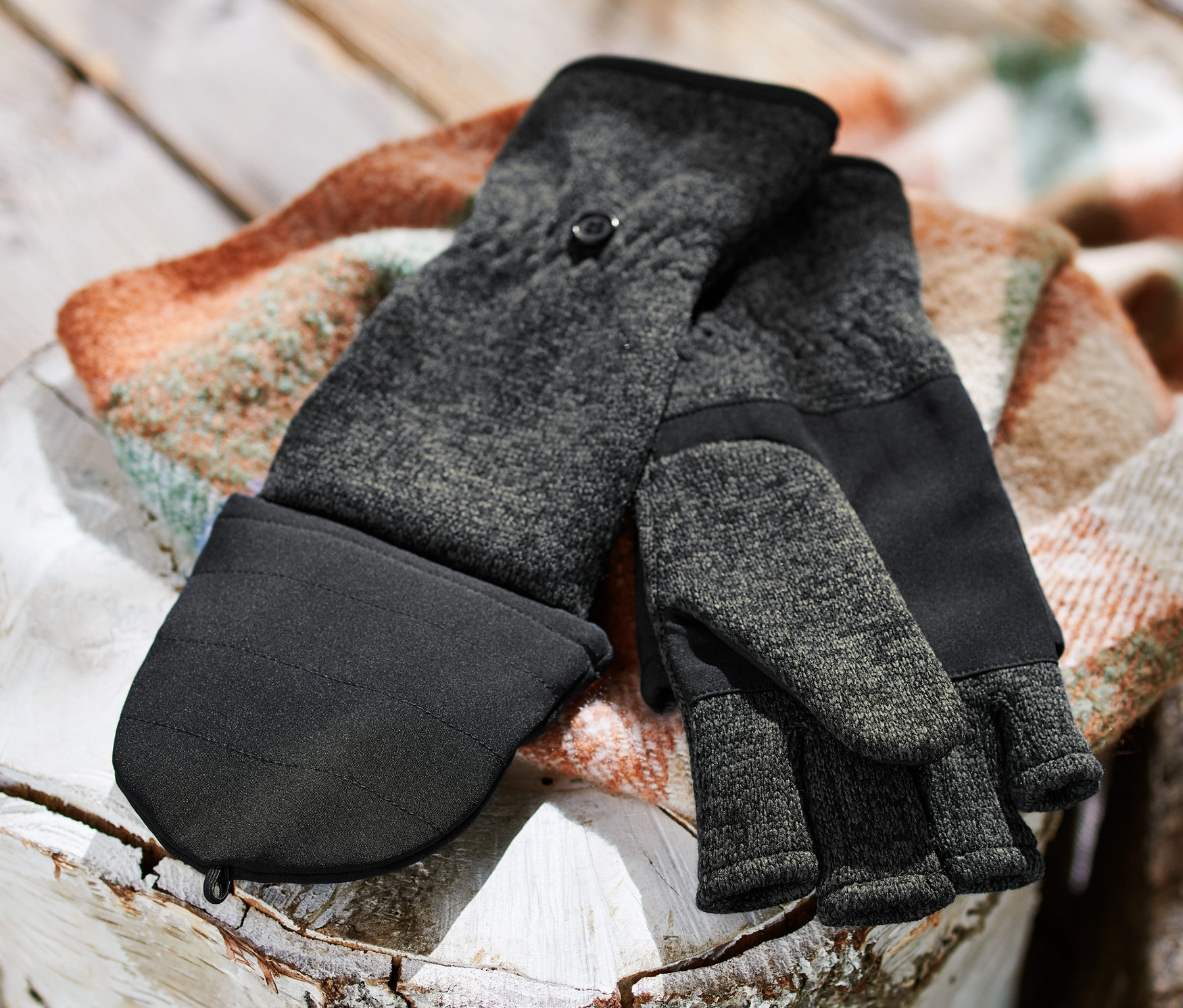 Ein Paar Strickfleece-Klappfäustlinge liegt auf einem Holzscheit neben einem Holzstapel.