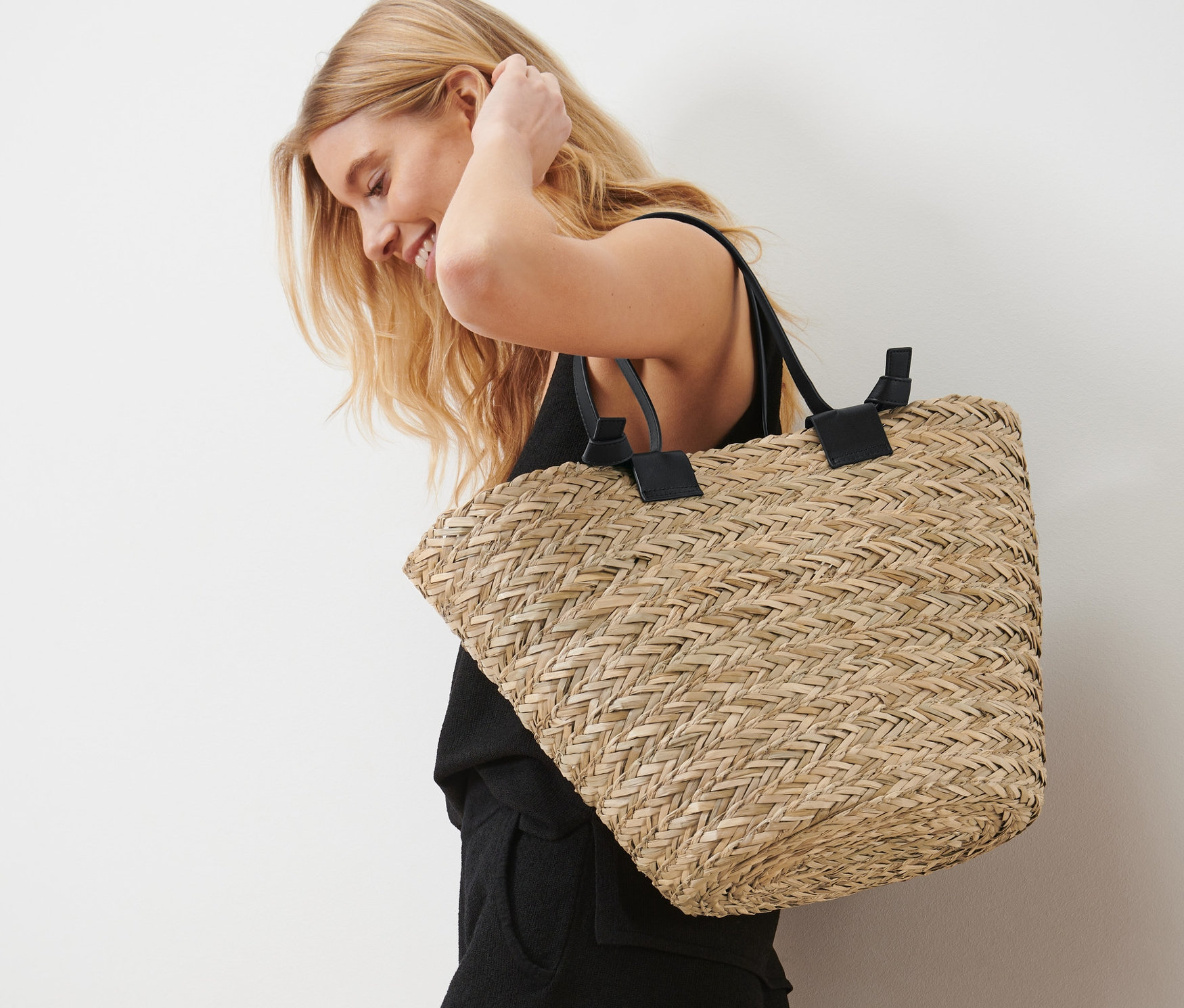 Blonde Frau lächelt und trägt eine Korbtasche über der Schulter.