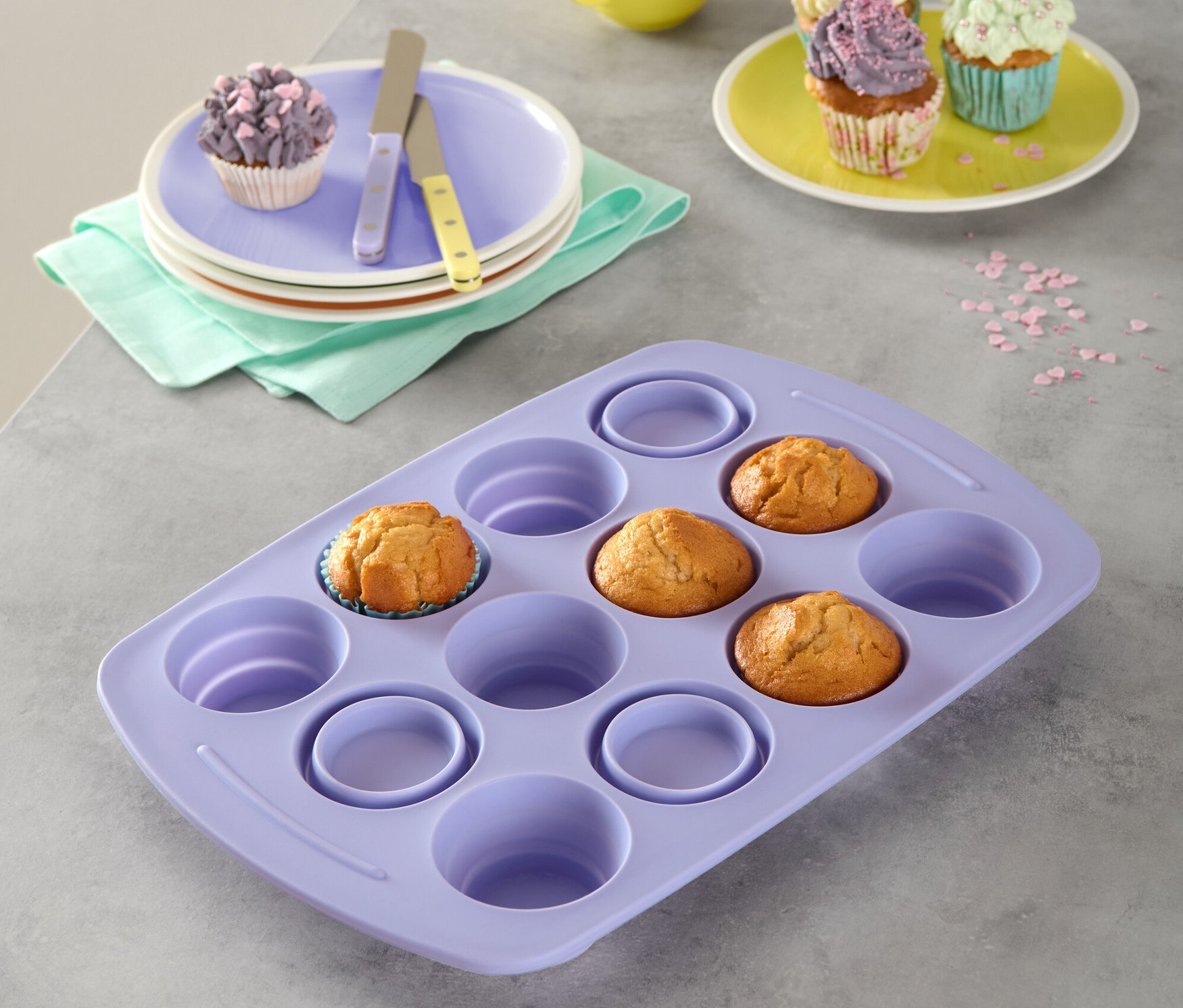 Lila faltbares Silikon-Muffinblech mit drei Muffins. Im Hintergrund Teller mit Muffins, gelber Becher und Streusel.