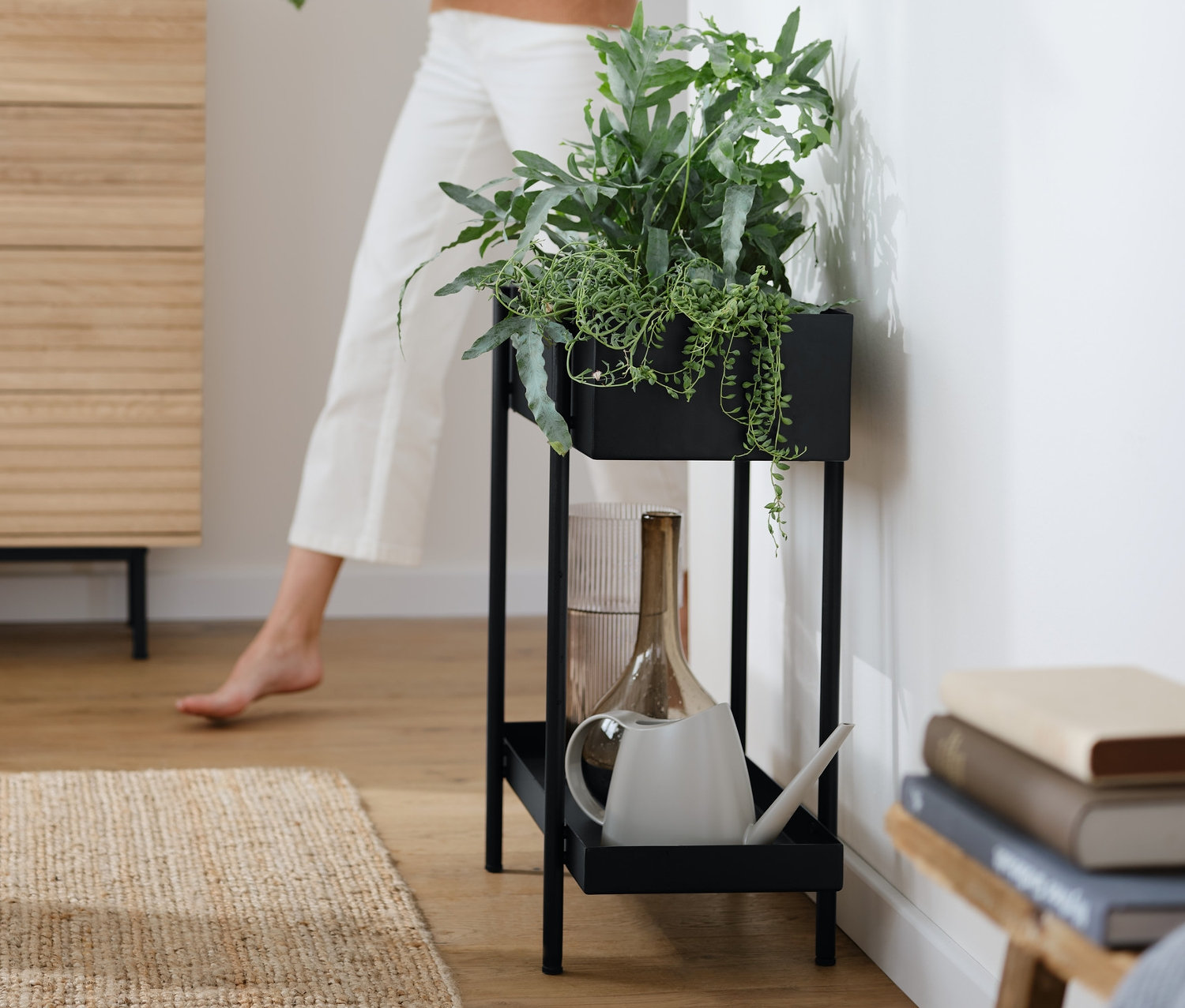 Eine Frau hält Pflanzen in der Nähe einer Kommode »Finnsbo« mit Lamellenoptik, auf der eine Tischleuchte mit Glaskugel, schwarz, und eine Plantbox »Skede«, groß, stehen.