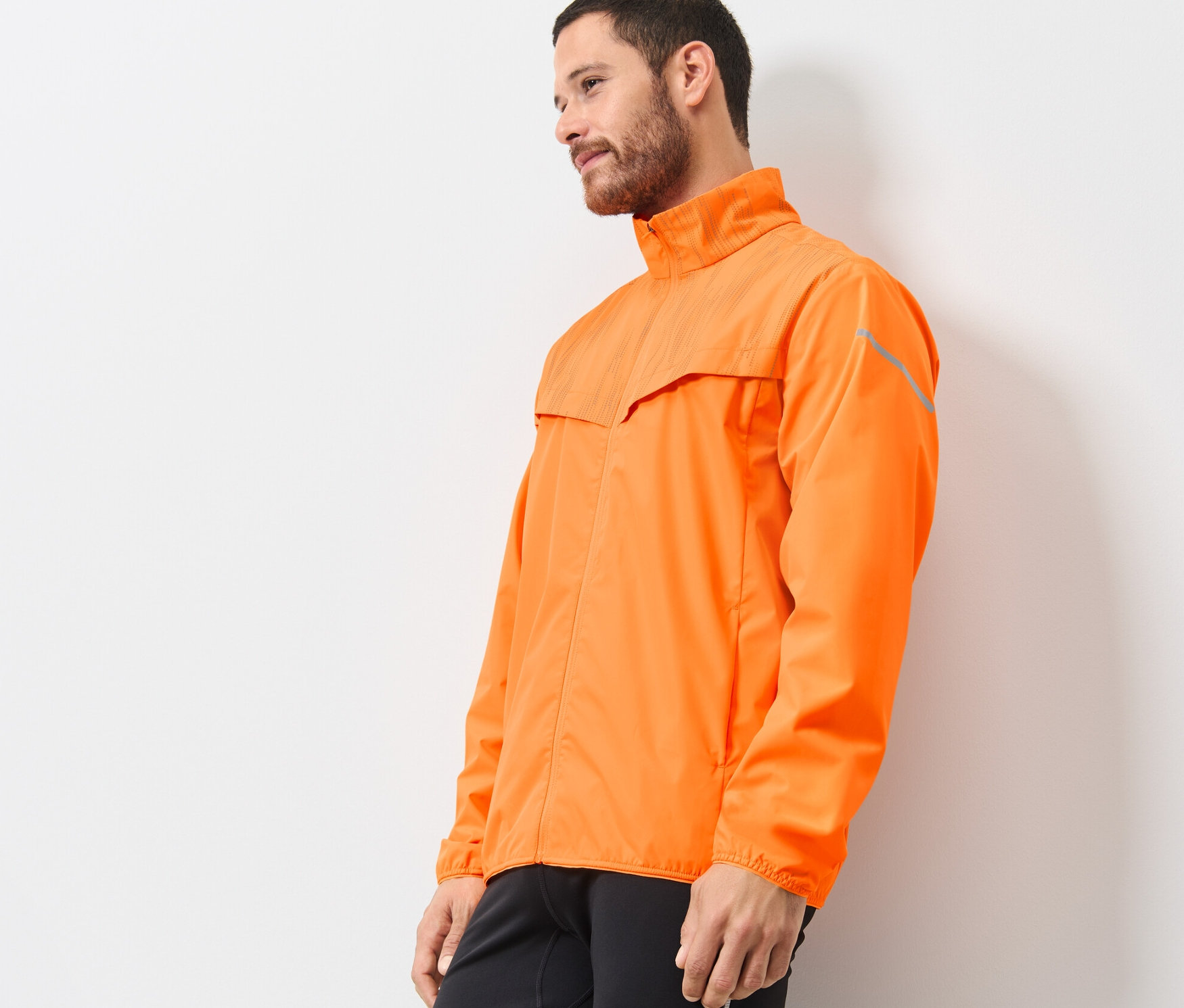 Mann trägt neonorange Windprotection-Laufjacke, seitlich stehend.