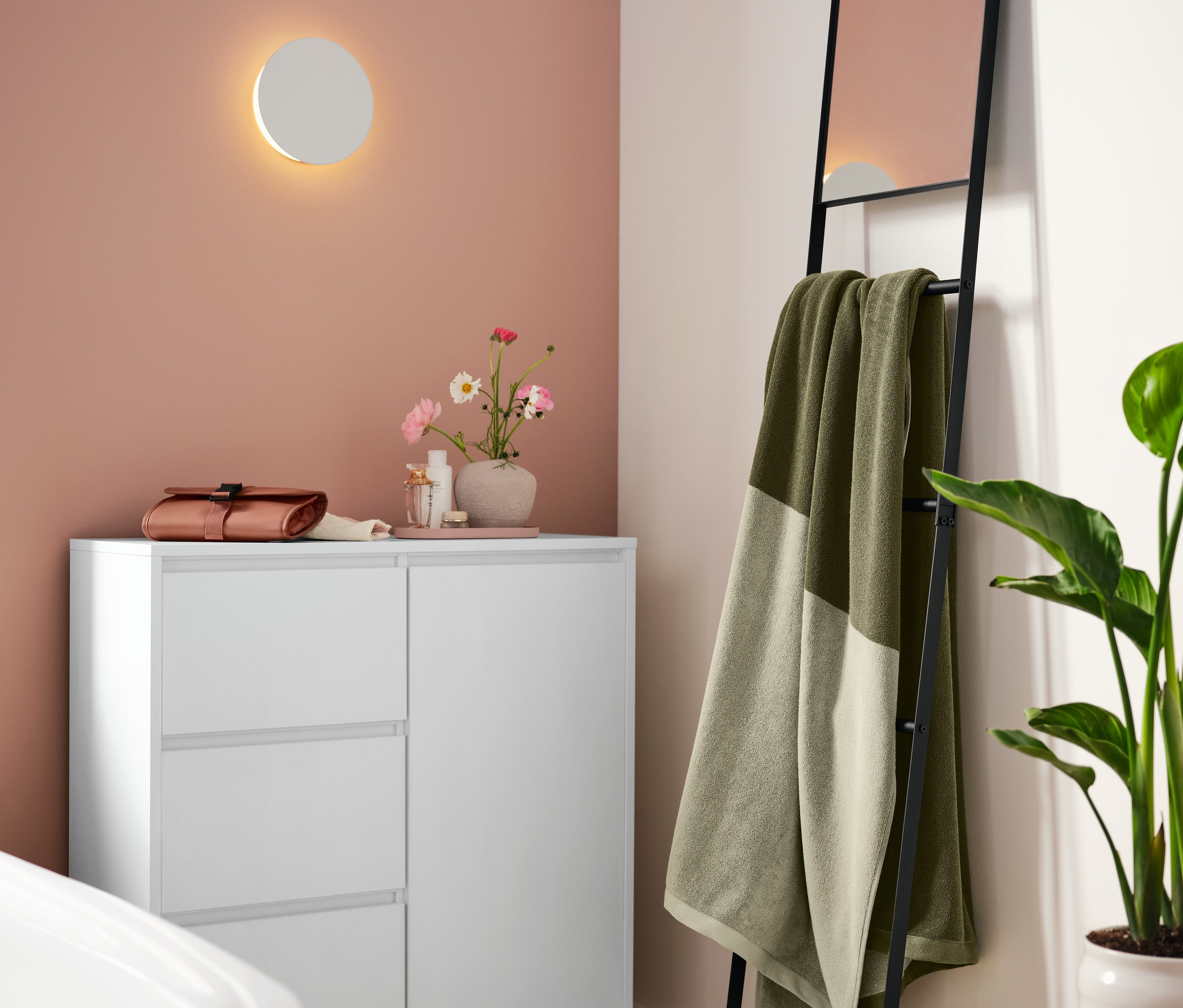 Im Badezimmer stehen ein weißes Bad-Sideboard, darauf ein rostfarbenes Tablett aus Metall und eine Kulturtasche. An der Wand hängt eine LED-Akku-Wandleuchte. An der Wand steht eine Handtuchleiter mit Spiegel, an der ein extragroßes grünes Badetuch hängt.