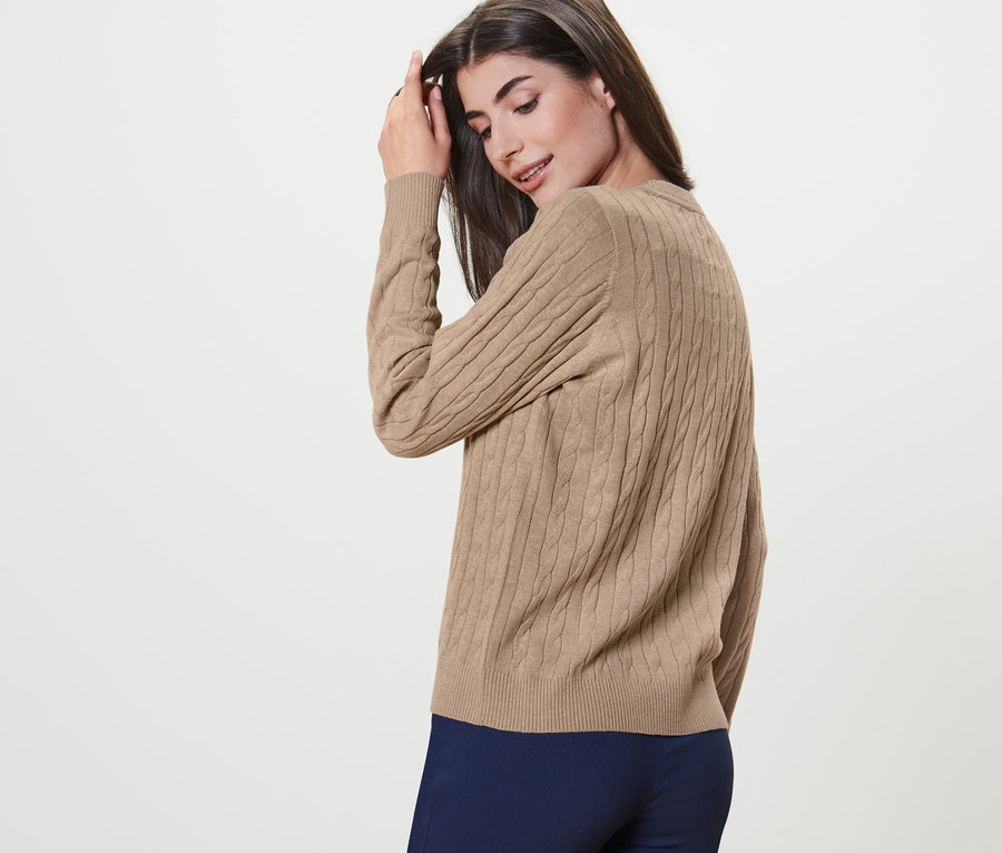 Frau posiert in einem creme melierten Zopfstrickpullover und dunkelblauen Hosen.