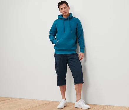 Mann lehnt an Wand, trägt blaugrünen Hoodie und navy Cargo-Bermuda in 3/4-Länge.