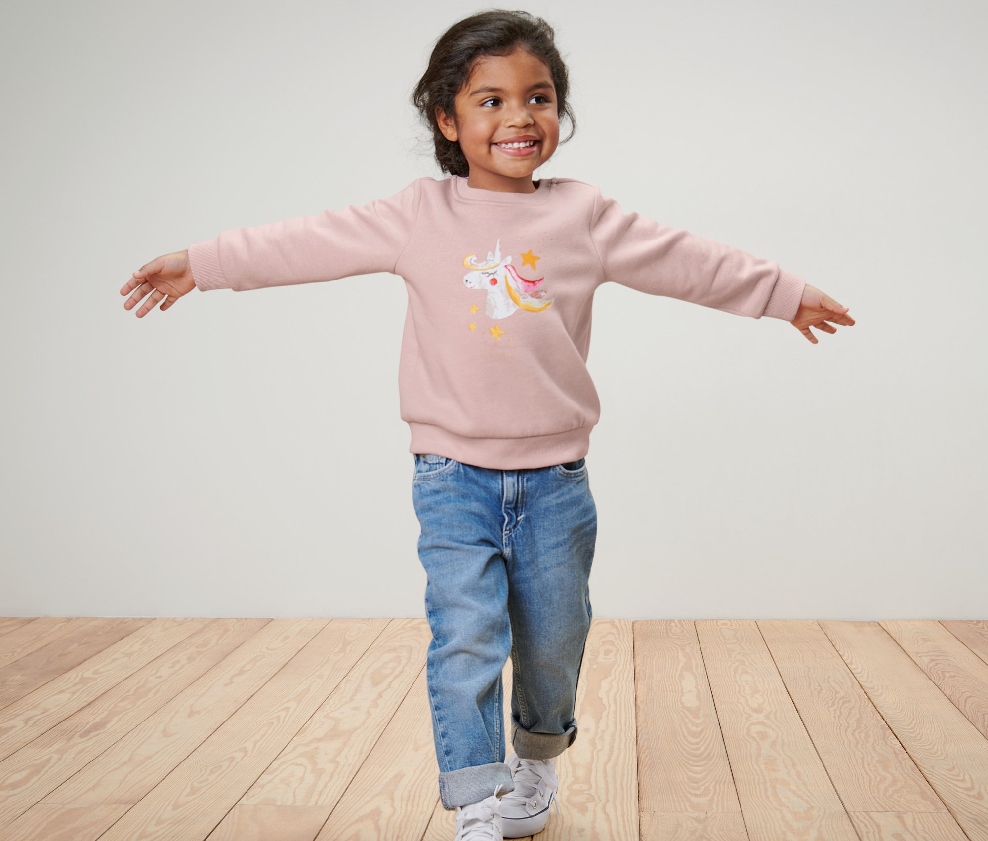 Mädchen mit ausgestreckten Armen trägt einen Kinder-Sweater mit Einhorn, blaue Jeans und Turnschuhe.