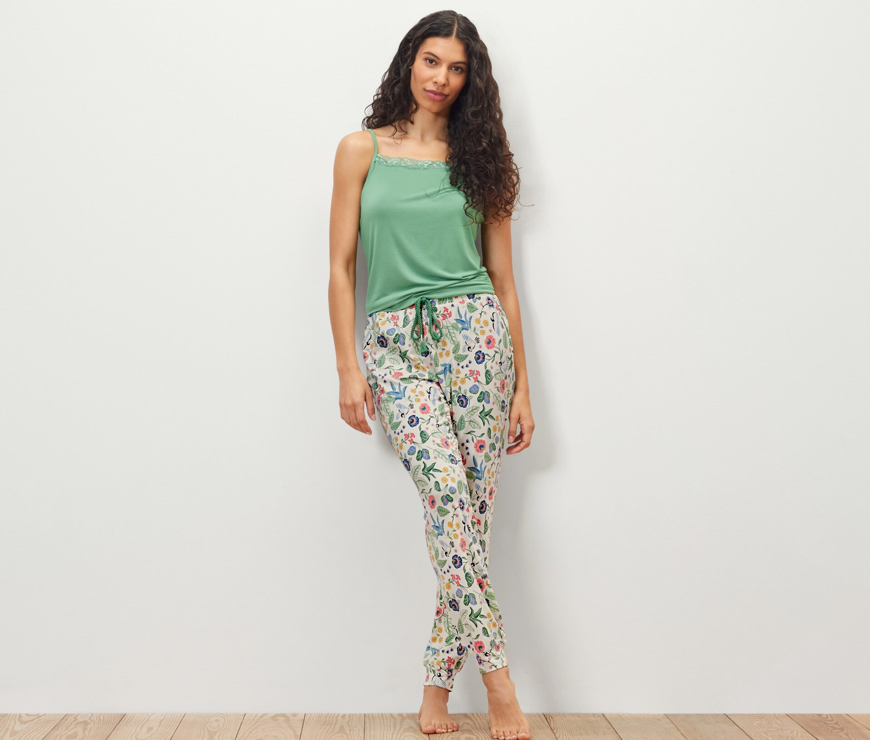 Eine Frau steht vor einer weißen Wand und trägt ein grünes Top und eine Pyjamahose.