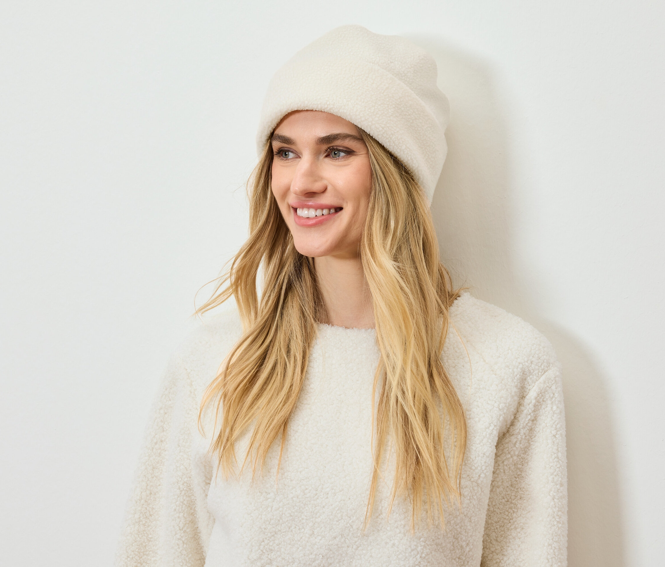 Lächelnde Frau mit blonden Haaren trägt eine cremefarbene Mütze und einen Pullover.