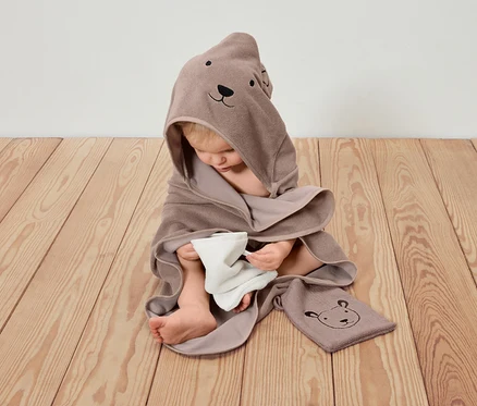 Ein Kind sitzt auf einem Holzboden, eingewickelt in ein braunes Kapuzenhandtuch mit Bärenmotiv und hält ein weißes Tuch.