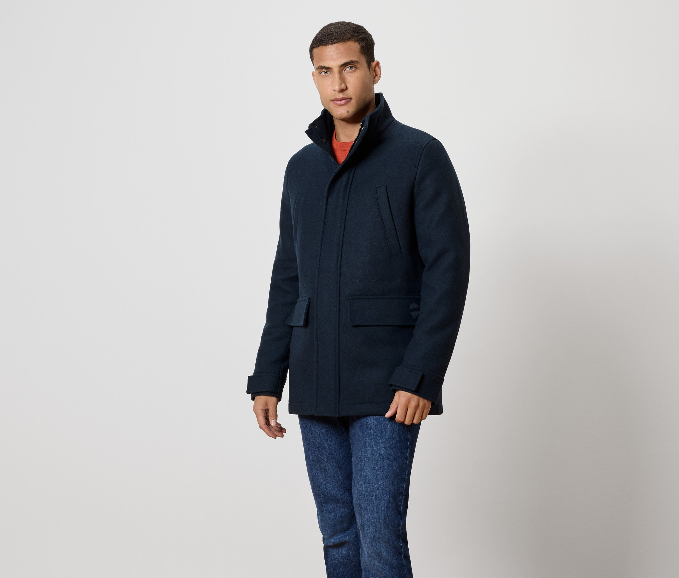 Mann in marineblauem Mantel und Jeans steht vor einem weißen Hintergrund.