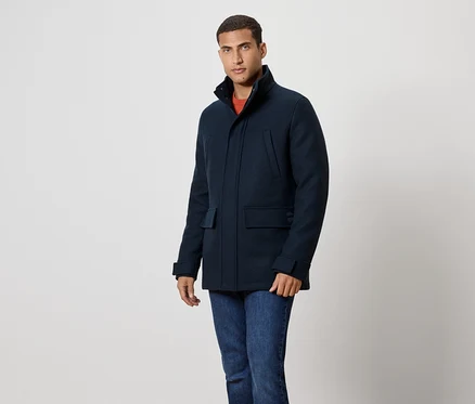 Mann in marineblauem Mantel und Jeans steht vor einem weißen Hintergrund.